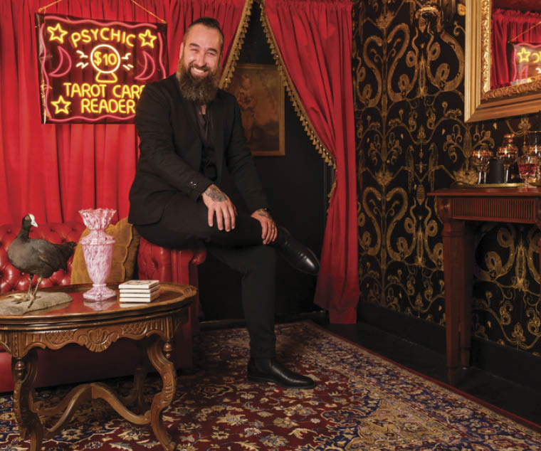 Homeowner Arryn poses in front of the converted casket lift bar cart in his former embalming room, now a converted speakeasy, as seen on HGTV’s We Bought a Funeral Home.